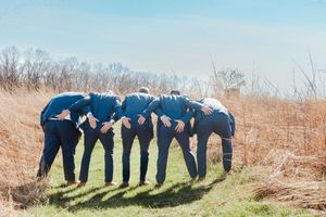 Group of men in blue suits huddled together in a circle on a grassy field with dry brush in background.