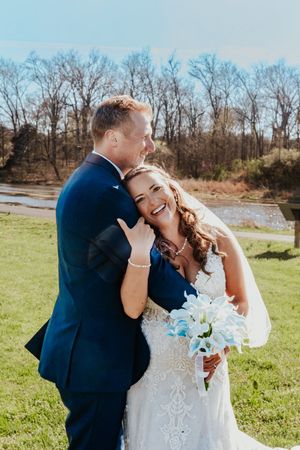 A joyful bride in a white lace gown holds blue flowers as a groom in a navy suit kisses her cheek outdoors.