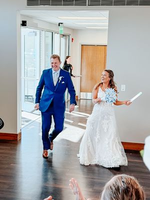 Bride in white lace gown and groom in blue suit make grand entrance at wedding reception, smiling joyfully.