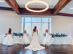 Bride in lace gown with three flower girls in white dresses standing in elegant wedding reception venue.