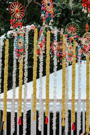 Colorful Indian wedding decor with marigold garlands, dream catchers, bells, and pompoms hanging outdoors.
