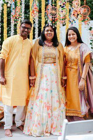Three people in traditional Indian attire smile together at a colorful mehndi celebration with decorative hangings.