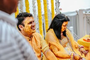 Couple laughing during Indian Haldi ceremony, groom in yellow kurta with turmeric on face, bride in traditional attire.