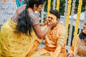 Couple celebrates Haldi ceremony in yellow outfits, with turmeric paste applied on groom's face during Indian wedding ritual.