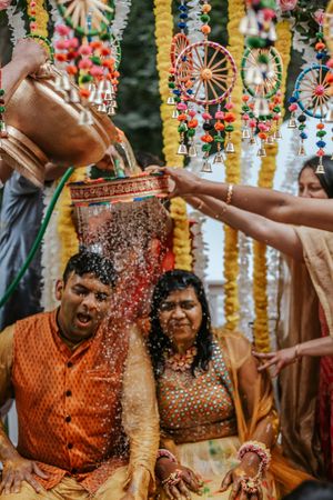 Groom in orange kurta gets water poured during a traditional Indian Haldi ceremony with colorful decorations.