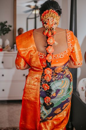Indian bride in vibrant orange silk saree with intricate floral hair braid adorned with marigolds, viewed from behind.