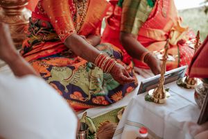 Indian bride in red saree with gold bangles sits cross-legged during traditional Hindu wedding ceremony with ritual items.