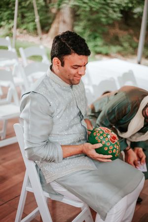 Man in traditional Indian attire holds decorative green floral pot during outdoor wedding ceremony.