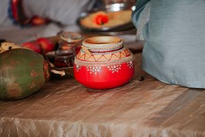 A vibrant red decorated Indian clay pot with white floral patterns sits on a table alongside a green coconut.