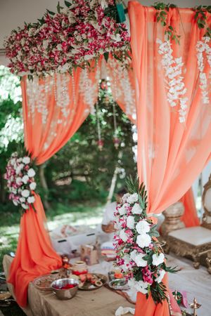 Elegant Indian wedding mandap decorated with coral orange drapes and pink white floral arrangements on a white draped table.