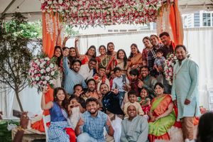 Large joyful group poses under floral mandap at a colorful traditional Indian wedding celebration.