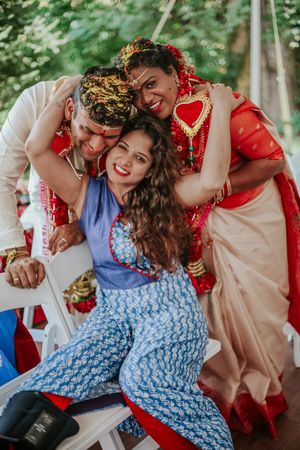 A joyful Indian wedding moment with a bride in red saree and groom embracing a smiling woman in blue floral outfit.