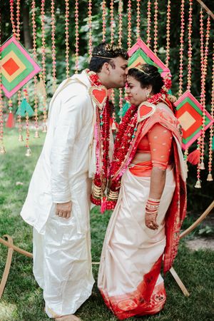 Indian bride and groom in traditional attire share a kiss at a colorful outdoor wedding ceremony with festive decorations.