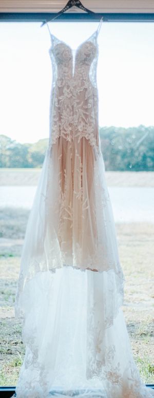 Elegant lace wedding dress with floral appliques and blush lining hanging by a window with scenic outdoor view.