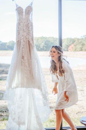 Excited bride in white robe admiring her lace wedding gown hanging by a sunlit window.