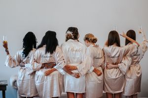 Six women in matching satin robes, including a bride, stand together arms around each other holding champagne glasses.