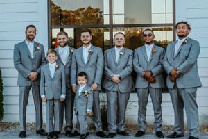 Groomsmen and groom pose in matching gray suits with boutonnieres outside a white barn wedding venue.