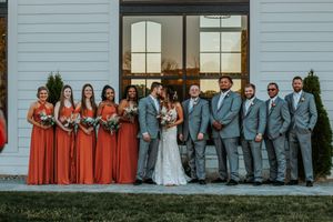 Wedding party posing outside white barn venue, bridesmaids in rust orange gowns and groomsmen in gray suits.