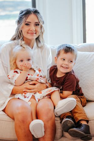 Smiling bride in white robe sitting on sofa with two young children during wedding morning preparations.