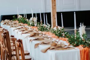 Elegant wedding reception head table with white candles, orange runner, and greenery floral arrangements.