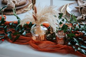 Rustic wedding tablescape with pampas grass in gold vase, greenery, and burnt orange cheesecloth runner.