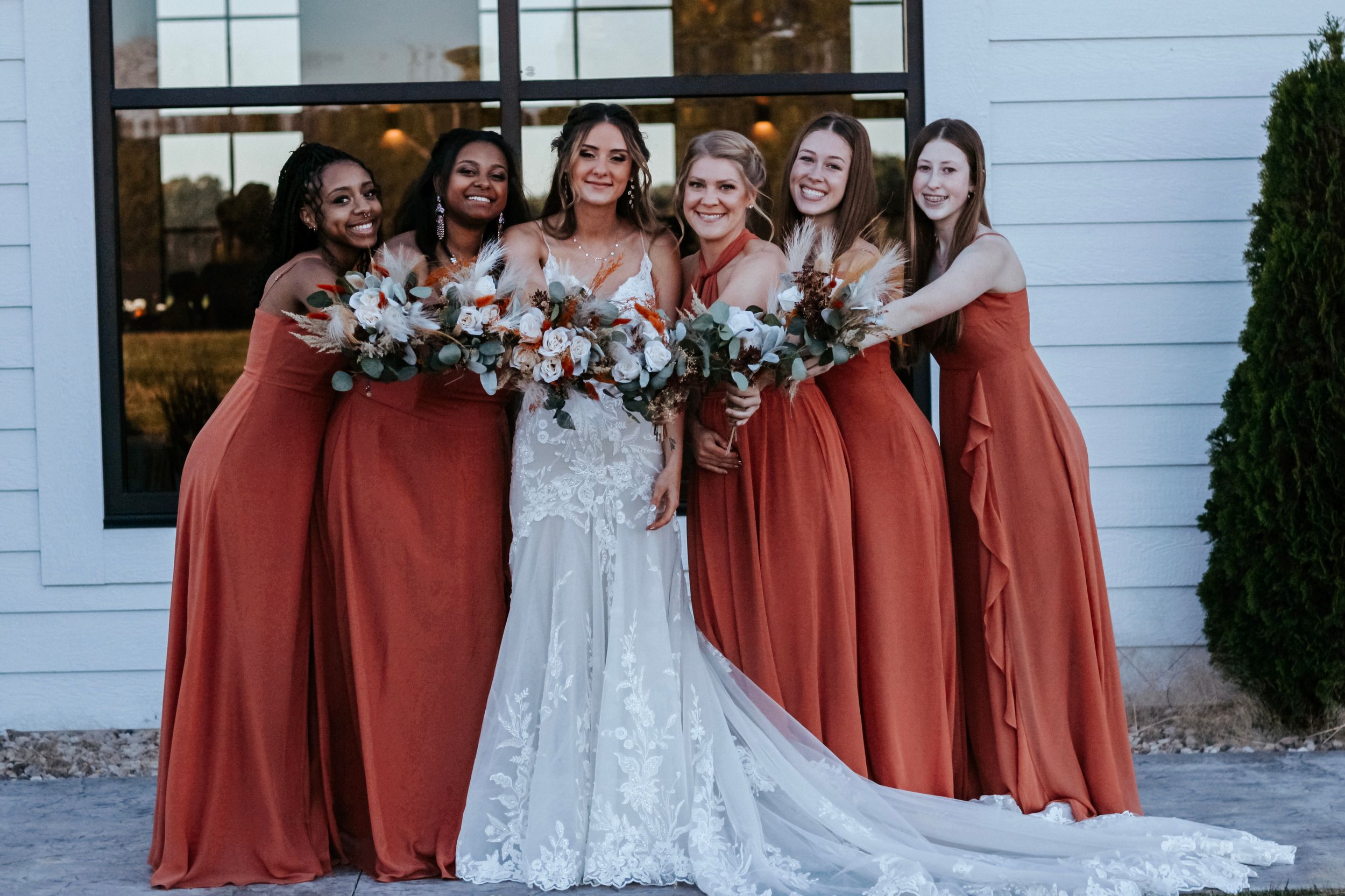 Bride in lace gown smiling with five bridesmaids wearing rust orange dresses holding bouquets outdoors.
