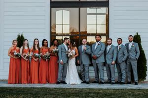 Wedding party poses outside white barn venue, bridesmaids in rust orange gowns and groomsmen in gray suits flanking bride and groom.