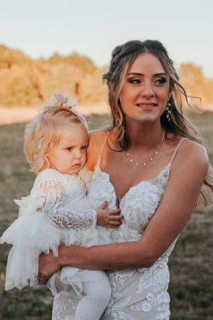 Bride in lace wedding gown holds a baby girl in white dress during golden hour outdoor wedding celebration.