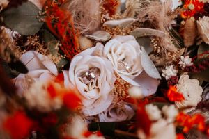 Close-up of wedding bouquet featuring white roses, orange dried flowers, and diamond engagement rings.
