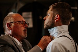 An older man adjusts the collar of a younger man in formal attire, likely a wedding preparation moment.