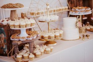 Elegant wedding dessert table featuring white tiered cake, cupcakes, cake pops, and gold accents on rustic wooden stands.