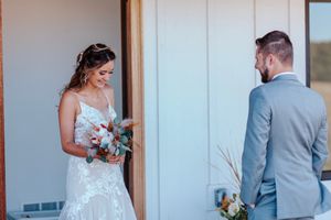 Bride in lace gown smiles holding bouquet during first look moment with groom in light gray suit at barn wedding.