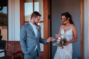 Groom in gray suit places ring on bride in lace wedding dress, holding bouquet, outside wooden doors.