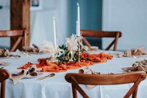 Elegant table setting with white candles, orange runner, dried florals, and wooden chairs in a bright dining room.