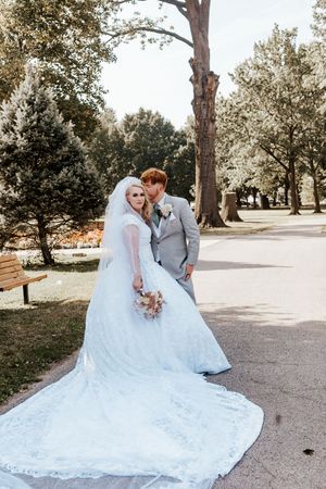 Bride in lace gown with veil and groom in gray suit pose on a park path surrounded by green trees.