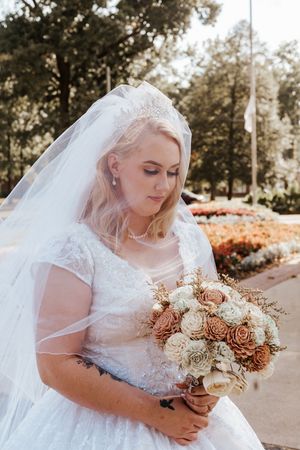 Blonde bride in white lace gown and veil holds bouquet of cream and terracotta roses outdoors.