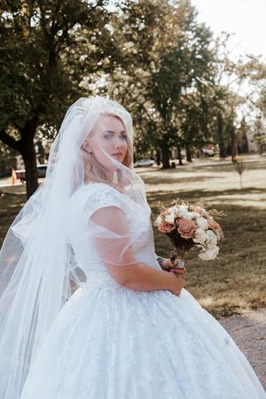 Blonde bride in white ballgown with veil holds bouquet in a sunlit park setting.