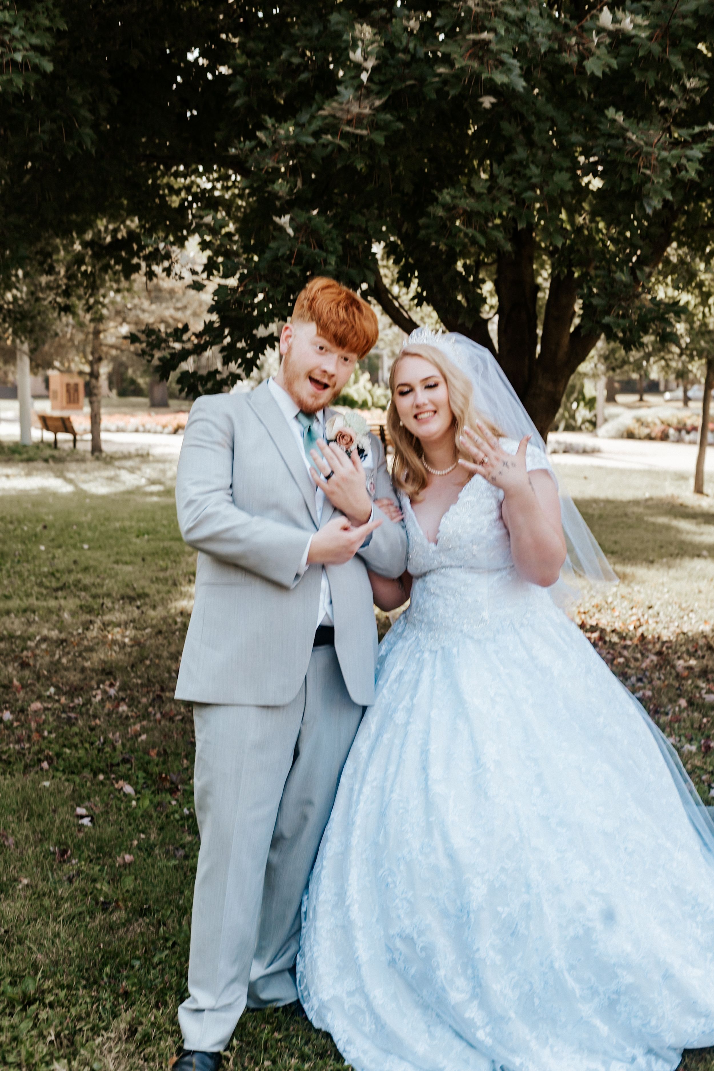 Newlywed couple in gray suit and blue lace ball gown showing wedding rings outdoors in a park setting.