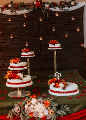 Elegant wedding cake display with red and white tiered cakes on gold stands, rose decor, and warm string lights backdrop.