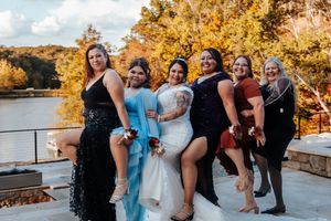 A bride in a white gown poses with five bridesmaids in colorful dresses by a scenic autumn lakeside.