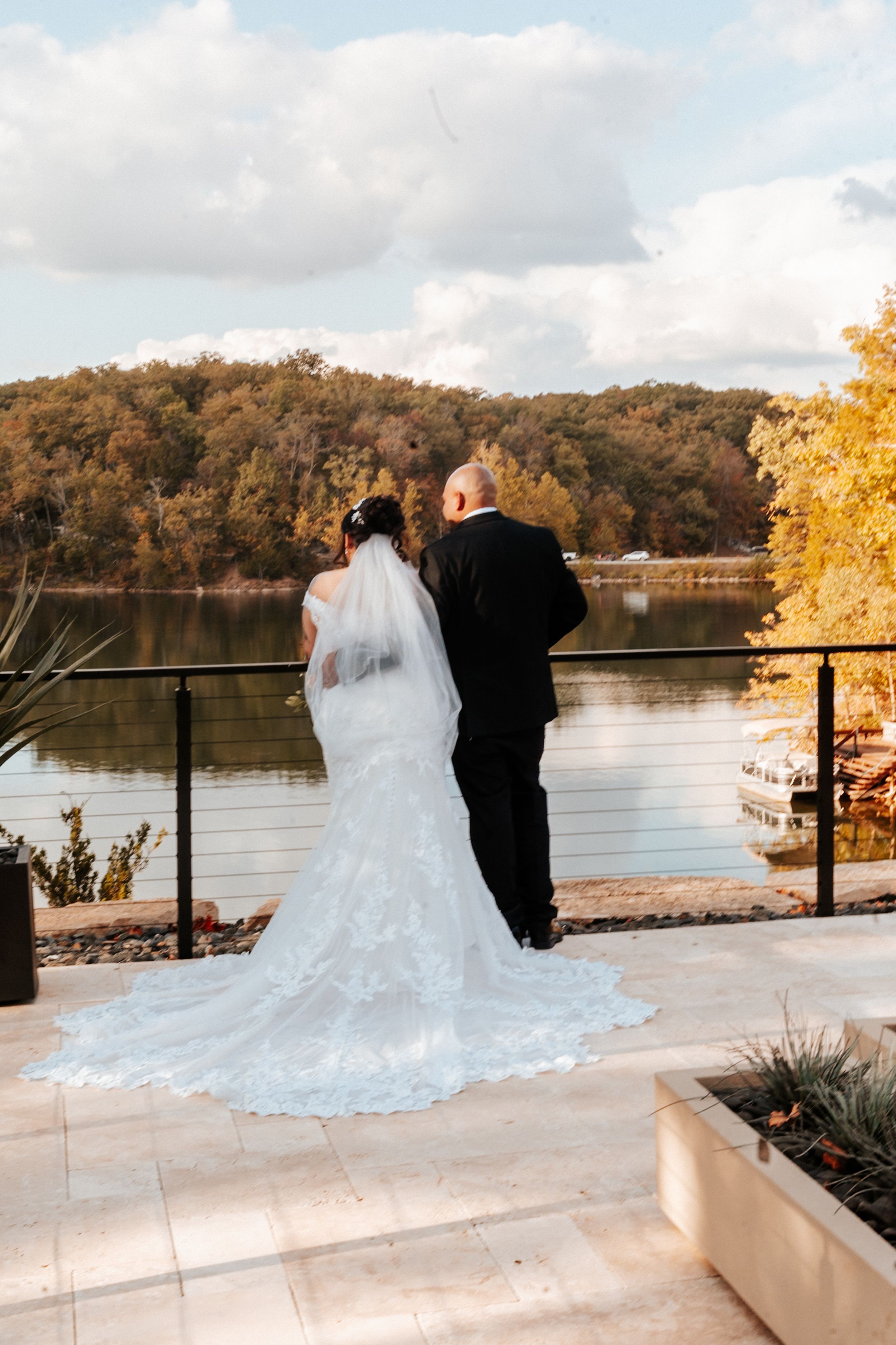 Bride and groom stand together on a lakeside terrace in autumn, bride wearing a lace mermaid gown with long veil.