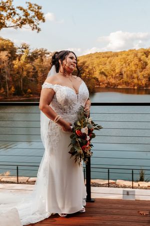 Bride in off-shoulder lace wedding gown holds bouquet on deck overlooking scenic autumn lake.