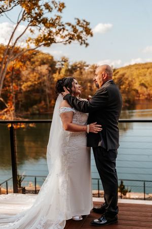 Bride and groom embrace on a deck overlooking a serene lake with colorful fall trees in the background.