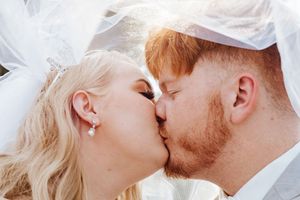 Newlywed couple sharing a romantic kiss under a bridal veil, bride wearing tiara and earrings, groom with red beard.