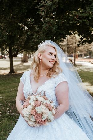 Blonde bride in white lace gown with veil and tiara holds blush bouquet outdoors.