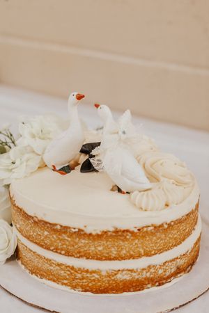 Elegant naked wedding cake with white frosting, decorative dove toppers, and white roses on a white surface.