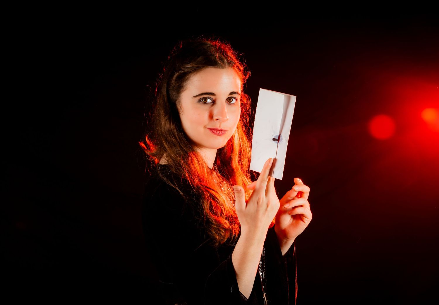 Young woman with red hair holding a white card against a dark background with red bokeh lights.