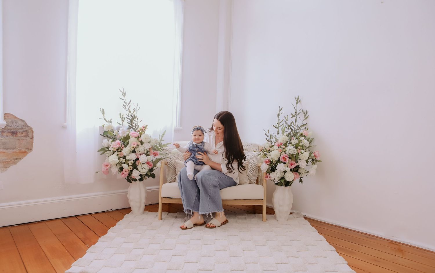 Mother holding baby on chair surrounded by floral arrangements in bright white room with hardwood floors.