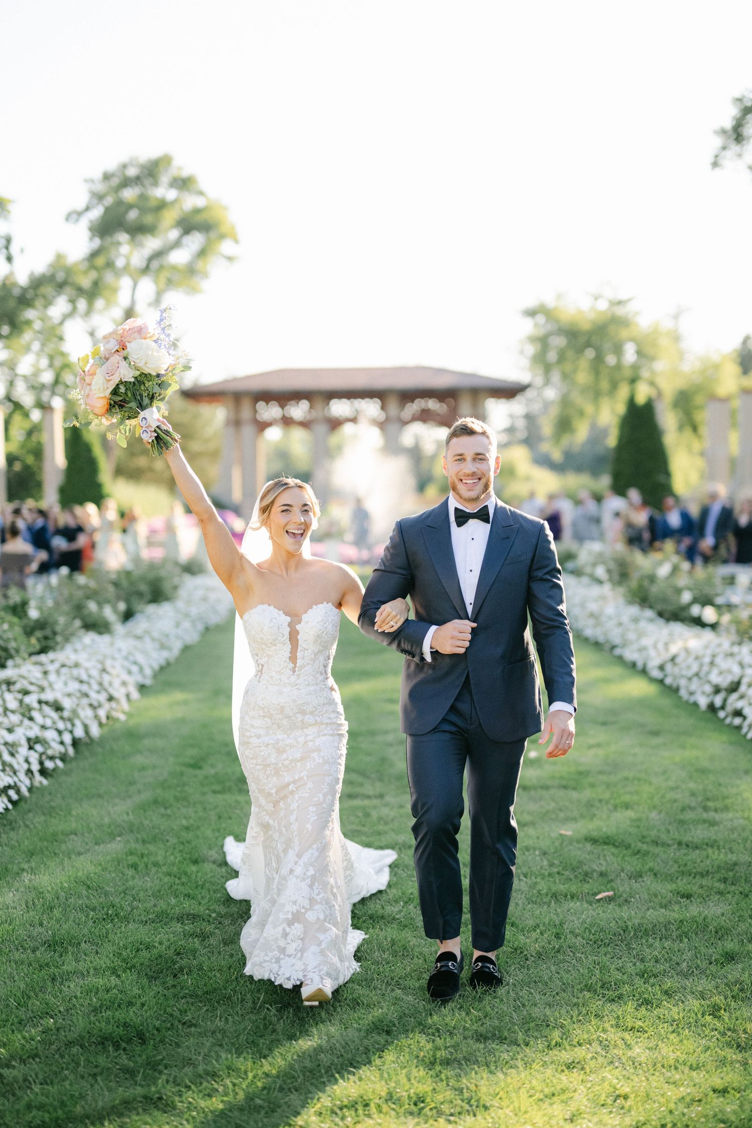 Armour House Wedding - Joyful bride in lace gown raises bouquet beside groom in navy tuxedo walking down outdoor wedding aisle.