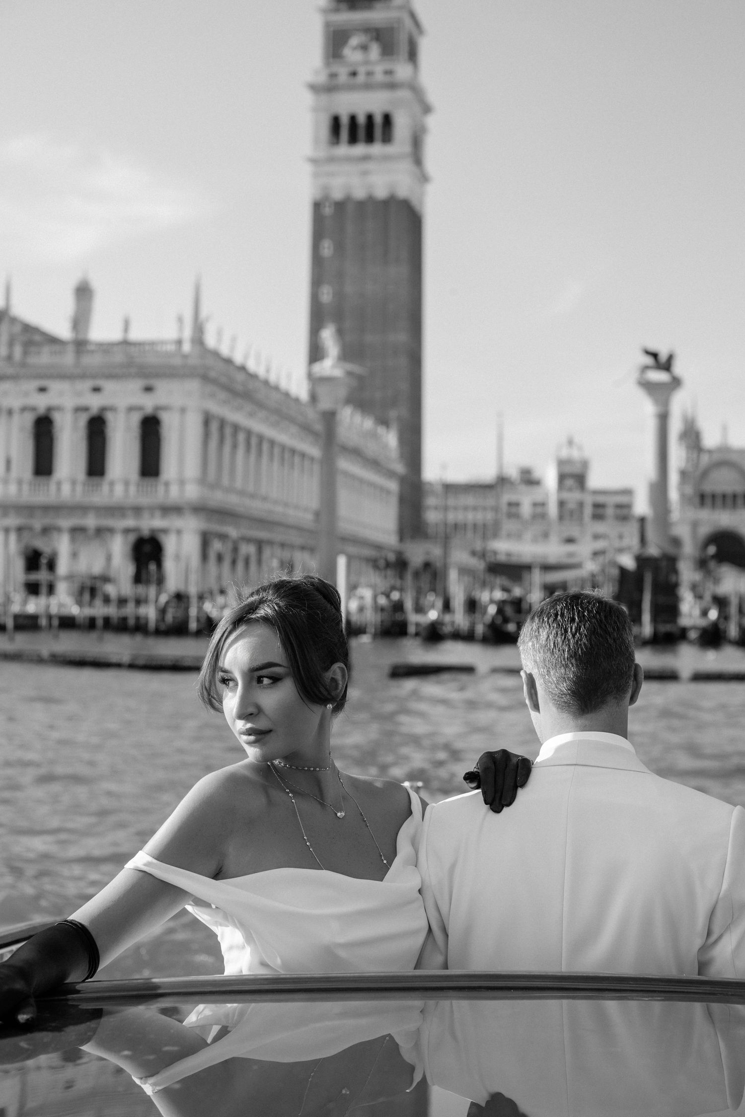 Couple in formal attire riding in a boat along Venice's Grand Canal with St Mark's Campanile tower in background.